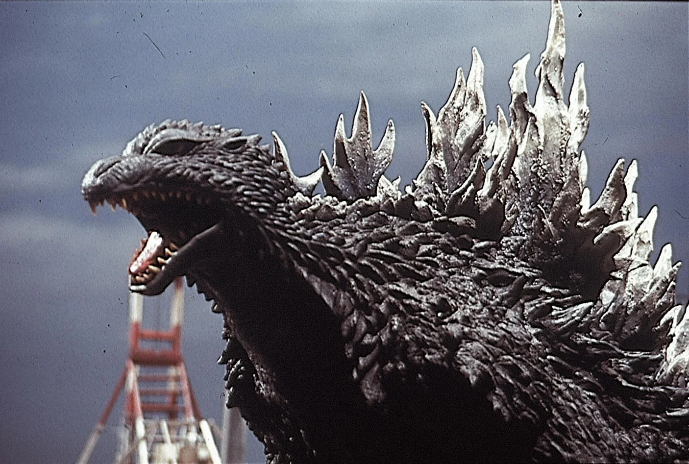 A profile shot of the Godzilla suit from Godzilla Against Mechagodzilla (2002): silver-tipped dorsal spines, mouth open mid-roar, with Tokyo Tower visible behind.