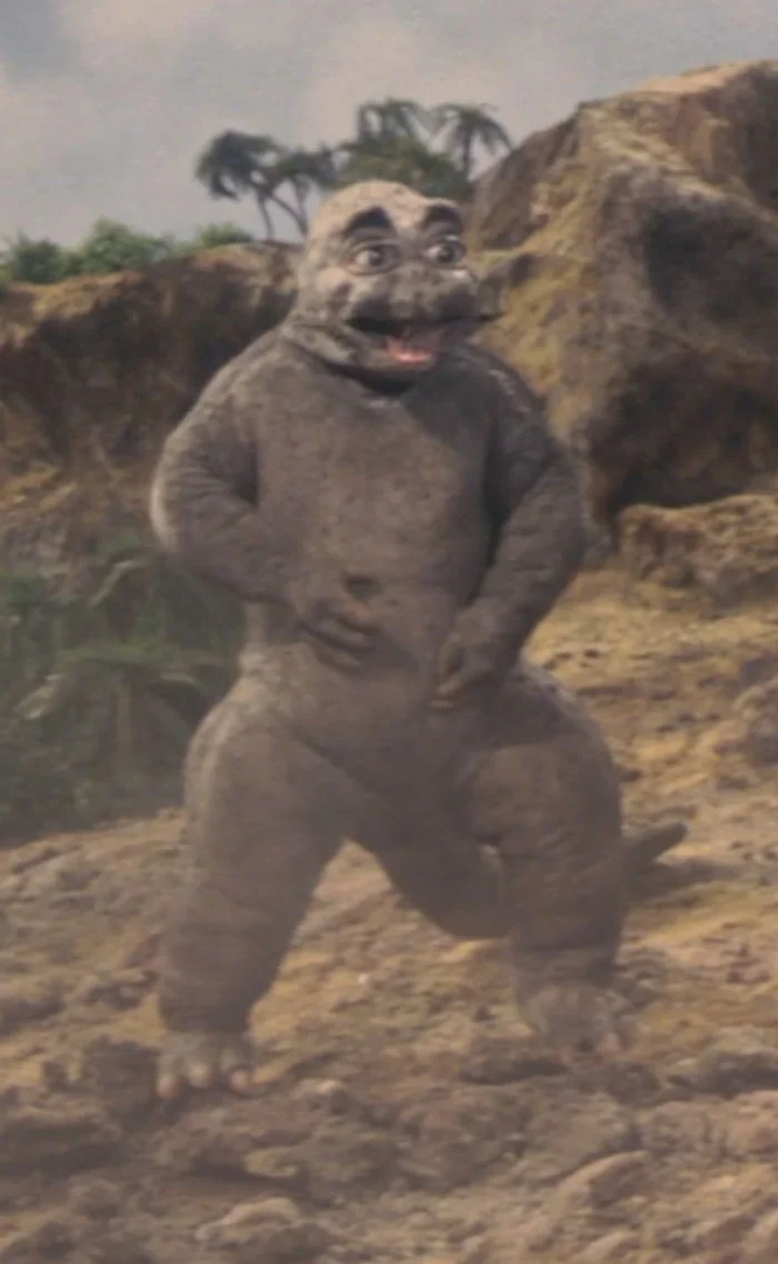 Minilla, the baby Godzilla from Son of Godzilla (1967): a small, round, gray creature with a friendly face, standing on a rocky island with palm trees in the background.