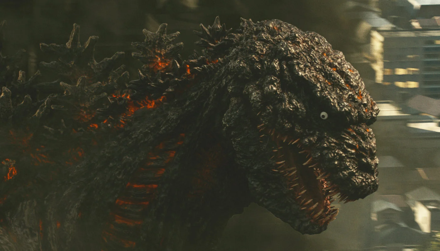 Close-up of the redesigned creature from Shin Godzilla (2016): bioluminescent red fissures across charcoal skin, vestigial forelimbs, and small lifeless eyes, framed against a blurred Tokyo skyline.