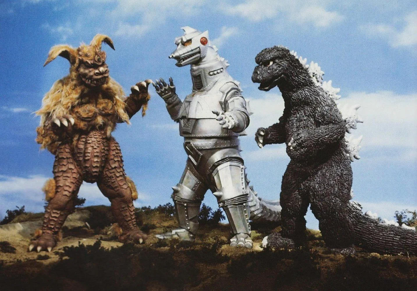 Three Showa-era kaiju on a grass-covered hillside under blue sky: King Caesar (the lion-like Okinawan guardian) on the left, Mechagodzilla in silver armor in the center, and Showa Godzilla on the right.